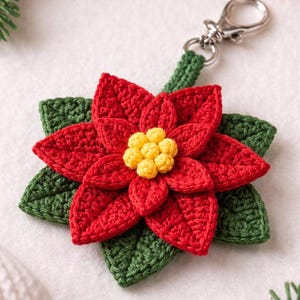 May include: A handmade crochet poinsettia keychain. The flower has vibrant red petals and a yellow center, with green leaves. It is attached to a silver-colored metal clasp and a green crocheted stem. The poinsettia is a festive holiday decoration.