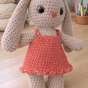 May include: A handmade crochet bunny doll in soft beige yarn, wearing a coral-coloured dress. The bunny has large black eyes and a small pink heart-shaped nose. The doll is approximately 30 cm tall.