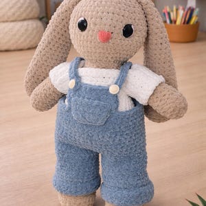 May include: A handmade crochet bunny doll with a tan body, long ears, and a pink nose. It wears a white shirt and blue overalls with button details. The plush toy is standing on a wooden surface.