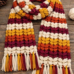 May include: A hand-knitted scarf in autumnal colours. The scarf has a textured pattern in cream, burgundy, orange, and yellow. The ends are finished with tassels in matching shades. A warm accessory for the colder months, perfect for adding a pop of colour.