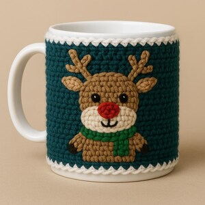 May include: A white ceramic mug with a teal crochet cosy featuring a reindeer design. The reindeer has brown antlers, a red nose, and a green scarf. The cosy is trimmed with white crochet.