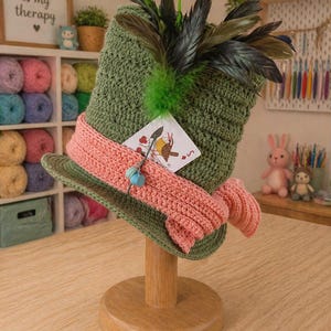 Peut inclure: Un chapeau vert crocheté à la main, orné d'une bande rose et de plumes. Il est présenté sur un support en bois. Une étiquette illustrée est attachée au chapeau. L'arrière-plan comprend de la laine, une pancarte "Crochet is my therapy" et des fournitures de loisirs créatifs.