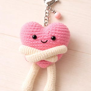 Puede incluir: Un llavero en forma de corazón rosa tejido a ganchillo con una cara sonriente, brazos y piernas color crema. El llavero tiene un cierre y una cadena plateados con un acento de cuentas rosas. El corazón se abraza a sí mismo.