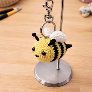 May include: A crocheted bee keyring. The bee is yellow with black stripes and white wings. It has small black eyes and a smiling mouth. The keyring is attached to a silver metal clasp and ring.