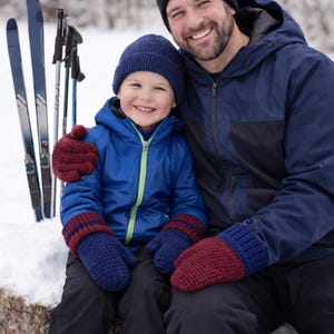 K&ouml;nnte beinhalten: Ein l&auml;chelnder Vater und ein Kind sitzen im Schnee. Das Kind tr&auml;gt eine blaue Jacke, M&uuml;tze und F&auml;ustlinge mit bordeauxfarbenen Streifen. Der Vater tr&auml;gt eine marineblaue Jacke und eine schwarze M&uuml;tze. Skiausr&uuml;stung im Hintergrund.