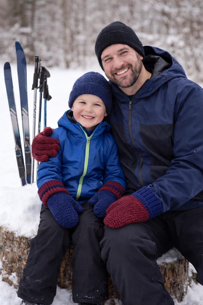 K&ouml;nnte beinhalten: Ein l&auml;chelnder Vater und ein Kind sitzen im Schnee. Das Kind tr&auml;gt eine blaue Jacke, M&uuml;tze und F&auml;ustlinge mit bordeauxfarbenen Streifen. Der Vater tr&auml;gt eine marineblaue Jacke und eine schwarze M&uuml;tze. Skiausr&uuml;stung im Hintergrund.