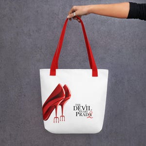 May include: White tote bag with red handles featuring a graphic of red high heels and the text "THE DEVIL WEARS PRADA". The bag is held up by a person.