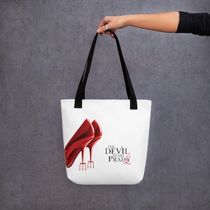 Könnte beinhalten: Weiße Tragetasche mit schwarzen Henkeln. Auf der Tasche sind rote High Heels mit Dreizack-Absätzen abgebildet. Der Text "THE DEVIL WEARS PRADA" ist in Schwarz und Rot aufgedruckt.