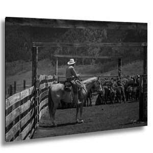 May include: A black and white photograph of a person wearing a cowboy hat, sitting on a horse inside a wooden corral. The person is facing a group of cattle standing in the dirt enclosure, with a mountainous landscape visible in the background.