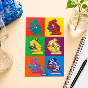 May include: A colorful pop art style notebook featuring six different colored microscope illustrations. The illustrations are in shades of green, red, yellow, purple, orange, and blue. The notebook has the text "IMMUNOgen" at the bottom.