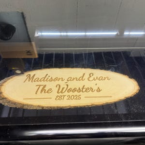 May include: A wooden oval sign with the text "Madison and Evan The Wooster's EST 2025" engraved in a light brown color. The sign has a natural wood grain edge.
