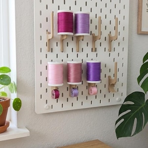 May include: A white pegboard displaying spools of thread in pink, purple, and magenta. The thread is held by wooden pegs. Black scissors lie on a wooden surface. A potted plant is on the left. The scene suggests a sewing or craft area.