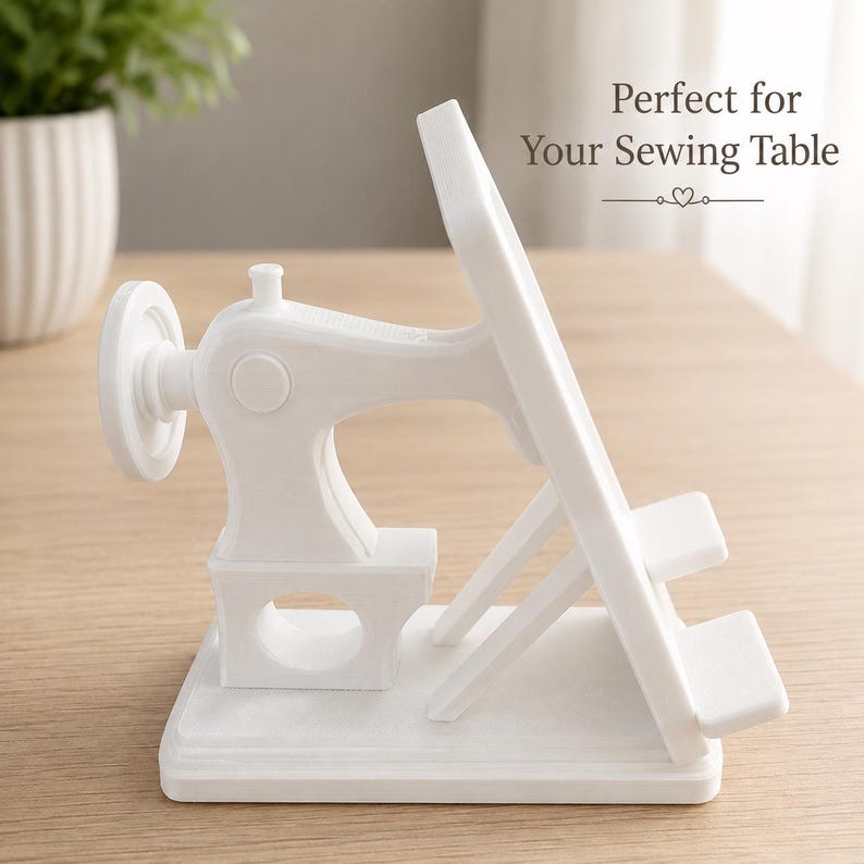 May include: A white plastic phone or tablet stand shaped like a vintage sewing machine, featuring a handwheel, a needle bar area, and a support ledge. The text "Perfect for Your Sewing Table" is printed above the stand with a small heart icon.