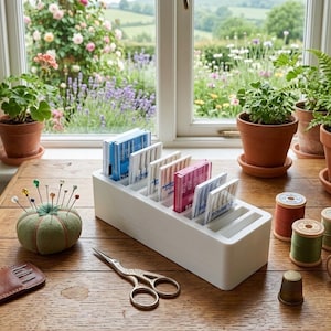 Può includere: Un organizer in plastica bianca contenente diverse custodie per aghi da macchina per cucire Schmetz di vari colori, un puntaspilli in tessuto verde con spilli colorati, forbici in metallo argentato, tre rocchetti di filo in legno e un ditale in ottone su un tavolo di legno.