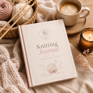 May include: A light pink "Knitting Journal" book with the text "The Makers Drawer" and illustrations of yarn and needles. The journal is next to a cup of coffee, a lit candle, and knitting supplies.