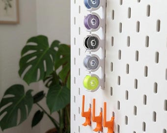 Puede incluir: Un panel perforado blanco con un soporte vertical para carretes que contiene cinco carretes de hilo en azul, morado, negro, gris y verde neón. Debajo hay tres ganchos naranjas y un recipiente de plástico transparente. Una planta en maceta está en el fondo.