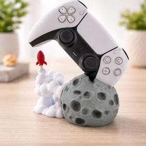 May include: A white and black video game controller rests on a gray moon-shaped base with a red rocket taking off. The controller has black buttons and a white surface. The rocket is red and the smoke is white.