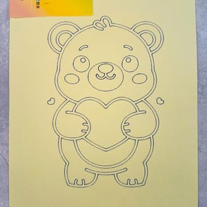 Peut inclure: Une feuille de papier jaune clair avec un dessin au trait noir d'un ours tenant un cœur. L'ours a de grands yeux, un petit nez et une bouche souriante. Le papier porte un logo dans le coin supérieur gauche qui dit "SANDRYLIX SAND PAINTING".