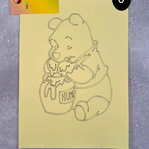 May include: A black outline drawing of Winnie the Pooh, seated and reaching into a honey pot. The honey pot has the word "HUM" on it. The drawing is on a yellow background. A business card with the name "SANDRYLIX" is in the upper left corner.