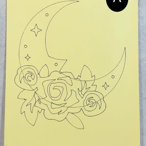 May include: A yellow sheet of paper featuring a black line drawing of a crescent moon, stars, and roses. The letter "A" is in a black circle. The drawing is centrally positioned on the paper.