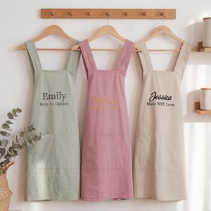 Puede incluir: Tres delantales en verde salvia, rosa empolvado y beige cuelgan de perchas de madera. El delantal verde salvia tiene el texto "Emily Born to Garden". El delantal rosa empolvado tiene el nombre "Yelicia". El delantal beige tiene el texto "Jessica Made With Love".
