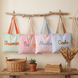 Puede incluir: Cuatro bolsas de mano personalizadas con nombres bordados en varios colores cuelgan de una rama de madera. Las bolsas son de colores pastel: verde menta, rosa, azul claro y azul pálido. Una cesta de mimbre, una suculenta en maceta y flores secas están sobre una mesa de madera.