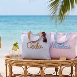Puede incluir: Dos bolsas de mano a rayas, una azul y otra rosa, sobre una mesa de mimbre cerca de la playa. La bolsa azul tiene bordado "Coda", y la rosa "Nicolas". Gafas de sol descansan sobre la bolsa azul.