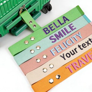 Puede incluir: Una maleta verde con una correa de equipaje verde a juego. La imagen muestra varias correas de equipaje coloridas con texto: BELLA, SMILE, FELICITY, Your text y TRAVER. Las correas son verdes, azules, rosas, beiges y naranjas.