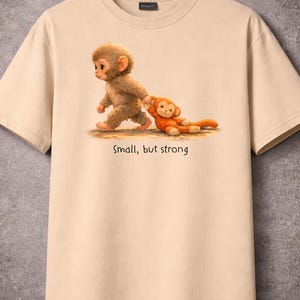 May include: Beige t-shirt featuring a charming illustration of two monkeys. One monkey is walking, pulling the other, which is lying down. The text "Small, but strong" is printed below the image. The shirt is hanging on a wooden hanger.