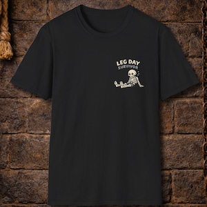 May include: Black t-shirt with a small graphic on the upper left chest. The graphic features a skeleton sitting down with the text "LEG DAY SURVIVOR" above it. The shirt has short sleeves and a crew neck.