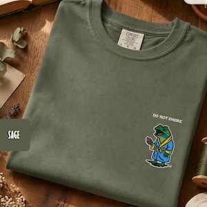 以下が含まれることがあります： セージグリーンのクルーネックTシャツ。青いバスローブを着てピンクの物体を持っている刺繍されたカエルが特徴で、カエルの上に「DO NOT SHIRE」の文字があります。Tシャツは木製の表面にあります。