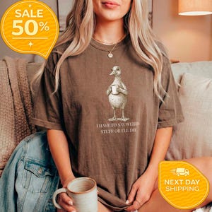 May include: A brown t-shirt featuring a whimsical illustration of a bird holding a mug, with the text "I HAVE TO SAY WEIRD STUFF OR I'LL DIE" below the image. The shirt has a relaxed fit.