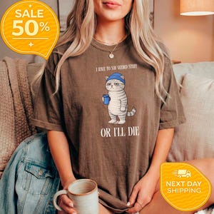 May include: Brown t-shirt with a graphic of a grumpy cat wearing a blue beanie and holding a blue cup. The text on the shirt reads "I HAVE TO SAY WEIRD STUFF OR I'LL DIE."
