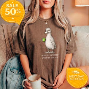 May include: Brown t-shirt featuring a quirky duck illustration holding a green mug. The text "I HAVE TO SAY WEIRD STUFF OR I'LL DIE" is below the duck. A yellow sale tag with "50%" is in the upper left corner.
