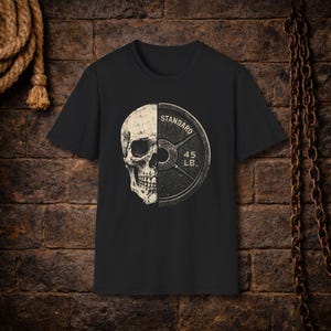 Może przedstawiać: Czarny t-shirt z grafiką przedstawiającą czaszkę połączoną z talerzem do podnoszenia ciężarów. Na talerzu widnieje napis "STANDARD" i "45 LB". Projekt wykorzystuje odcienie bieli i szarości, kontrastujące z czernią koszulki.