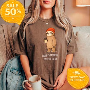 May include: Brown t-shirt featuring a cartoon sloth wearing an orange beanie and holding a coffee cup. The text "I HAVE TO SAY WEIRD STUFF OR I'LL DIE" is printed below the sloth. A yellow sale tag is in the upper left corner.