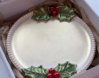May include: A round, cream-coloured ceramic plate with a beaded edge, featuring two green holly leaf accents with three red berries each at the top and bottom. The plate is nestled in a white box with brown tissue paper and a label reading Maleta Candles.