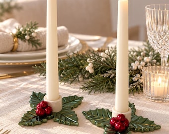 May include: Two off-white taper candles standing in ceramic candle holders shaped like green holly leaves with clusters of three glossy red berries. The candles are lit, casting a warm glow, and the holders are placed on a textured fabric surface.