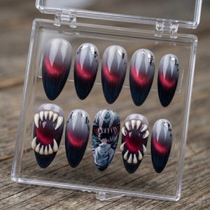 May include: A set of ten almond-shaped press-on nails in a clear plastic case. The nails feature a dark gray to black gradient with a red cat-eye effect and silver foil accents. Two nails display 3D white fangs, and one features an anime character.