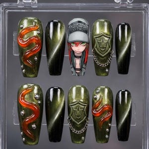 Puede incluir: Un juego de diez uñas postizas en una caja de plástico transparente. Las uñas presentan una variedad de diseños, incluyendo una cara de dibujos animados, un escudo con una cadena y un diseño de serpiente roja sobre fondo verde.