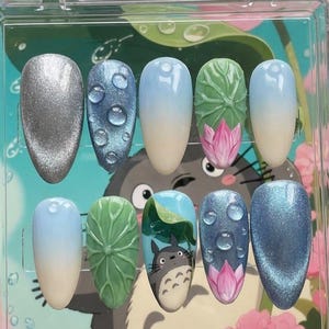 Op de afbeelding: Een set van tien kunstnagels in een doorzichtige plastic doos. De nagels hebben verschillende ontwerpen, waaronder waterdruppels, een Totoro-illustratie en lotusbloemen. Kleuren zijn onder meer zilver, blauw, groen en roze.