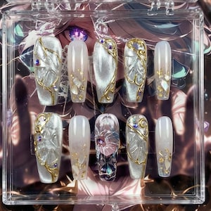 May include: A set of ten artificial nails in a clear plastic case. The nails feature a variety of designs, including silver and gold accents, gemstones, and intricate patterns. Some nails have a clear base with gold flakes.