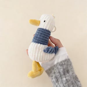 Könnte beinhalten: Ein Plüsch-Entenspielzeug mit weißem Körper, blauem Pullover und gelbem Schnabel und Füßen. Die Ente hat eine Cordstruktur und eine kleine Schlaufe oben. Das Spielzeug wird in einer Hand gehalten.