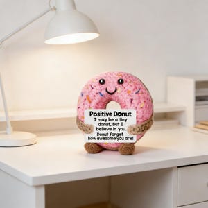 May include: A pink crocheted donut with sprinkles, holding a sign that reads "Positive Donut." The sign has a motivational message. The donut has a smiling face and is on a white desk with a lamp.