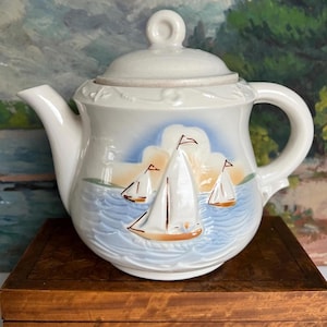 May include: White ceramic teapot with a sailboat design. The teapot features a lid and handle, with a painted scene of sailboats on blue water under a light blue sky. The teapot is sitting on a wooden box.