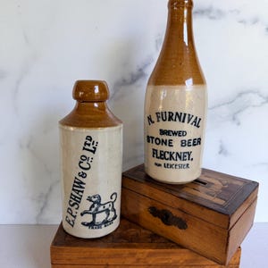 May include: Two vintage stoneware bottles with brown glazed tops and cream-colored bodies. One bottle features the text E.P. SHAW & CO LTD and a dog graphic. The other bottle features the text N. FURNIVAL BREWED STONE BEER FLECKNEY FOR LEICESTER.