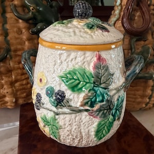 May include: A ceramic jar with a textured cream-colored surface, featuring raised relief designs of green leaves, yellow flowers, and dark purple blackberries. The lid is topped with a blackberry finial, and the jar has two dark green handles.