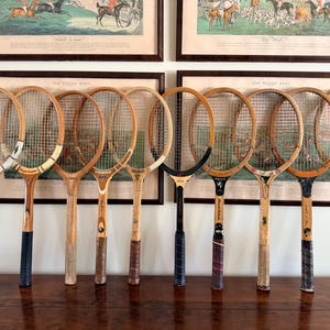 May include: A collection of nine vintage wooden tennis rackets arranged in a row. Each racket features a distinct wooden frame, nylon stringing, and a handle wrapped in various colored grip tapes, including white, black, and brown leather textures.