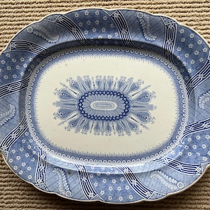 May include: Oval serving platter with a blue and white floral design. The platter has a scalloped edge and a central oval motif. The border features a repeating pattern of flowers and geometric shapes. A classic piece for serving food.