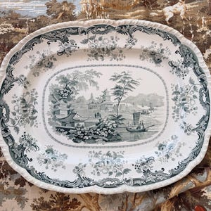 May include: An oval ceramic serving platter featuring a dark green transferware pattern on a white background. The central scene depicts a landscape with pagodas, trees, and boats on water, surrounded by a decorative floral border with scalloped edges.